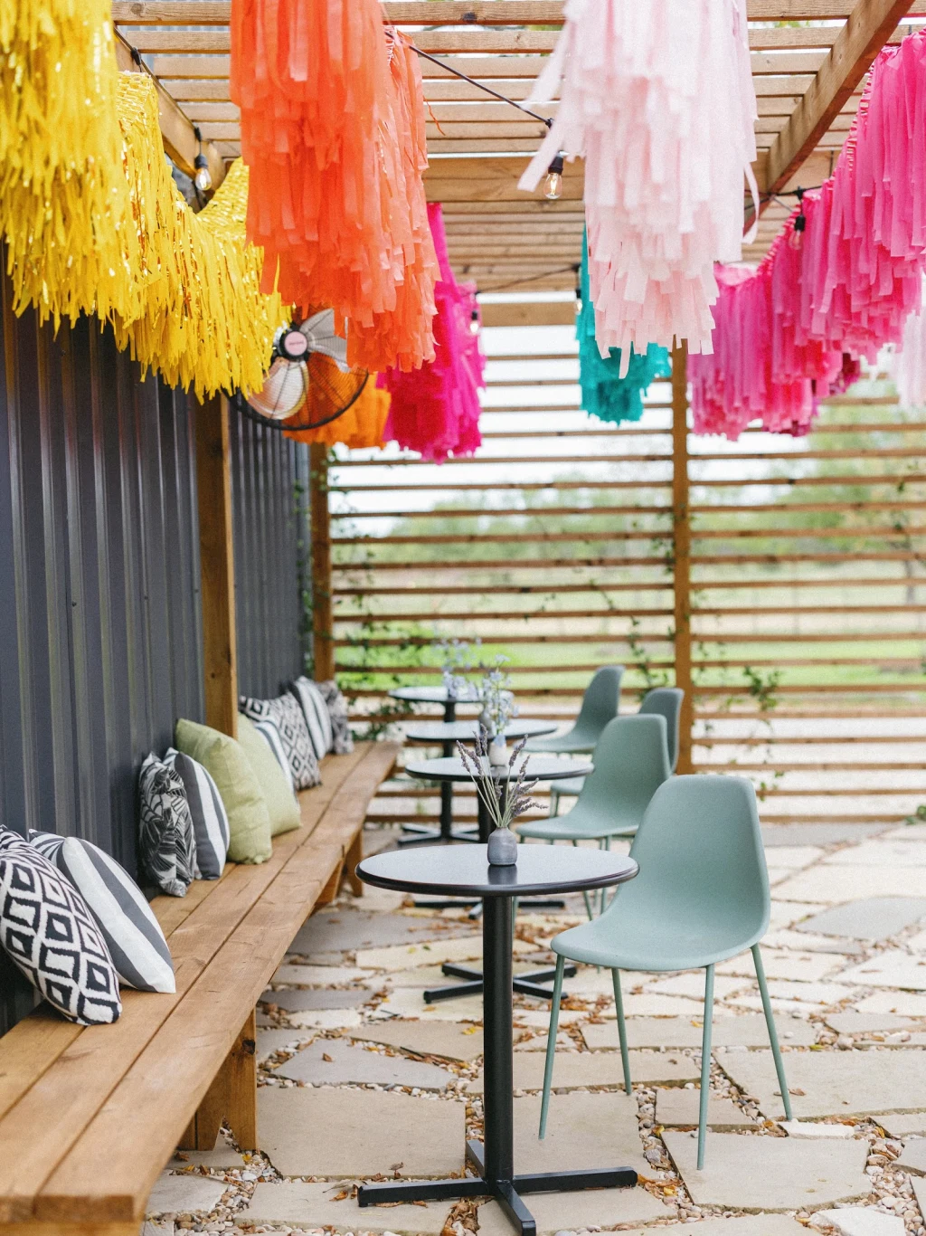 Covered cedar patio at Rancho Moonrise set with round bistro tables, sage green chairs, and a long bench lined with patterned pillows, paper fringe garlands and string lights overhead