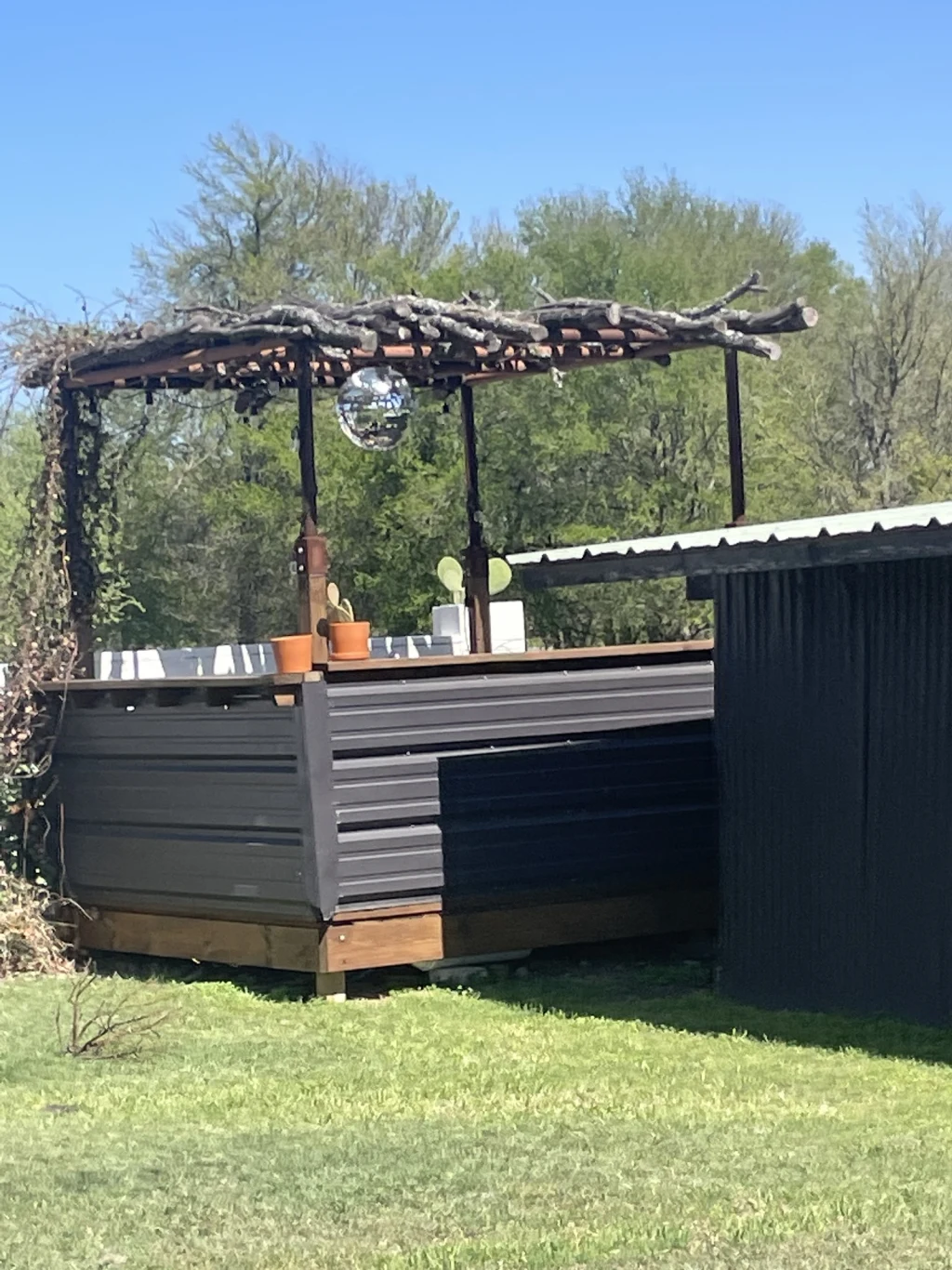 Outdoor event bar at Rancho Moonrise clad in corrugated metal with a wood counter, potted prickly pear, and a disco ball hanging from a pergola built of raw cut branches