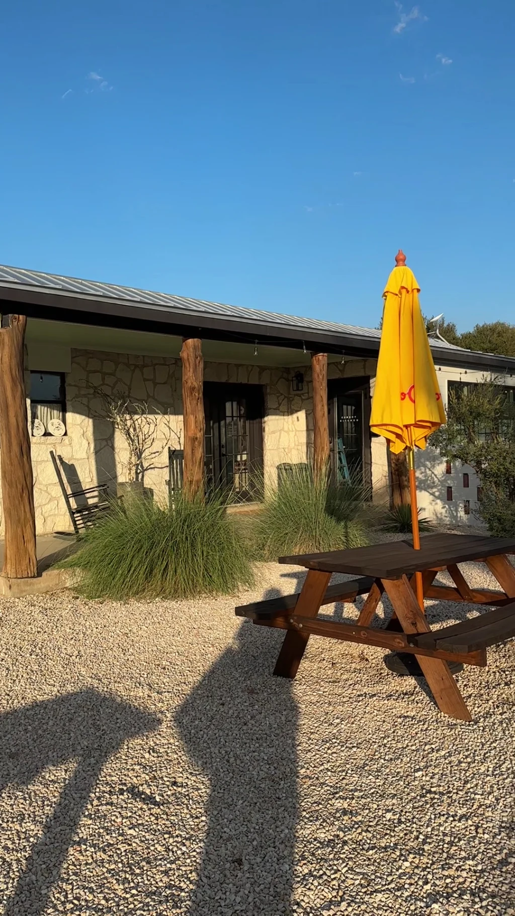 Stone-faced Lodge porch at Rancho Moonrise with a wood picnic table, open yellow umbrella, rocking chairs, and cedar log posts at golden hour