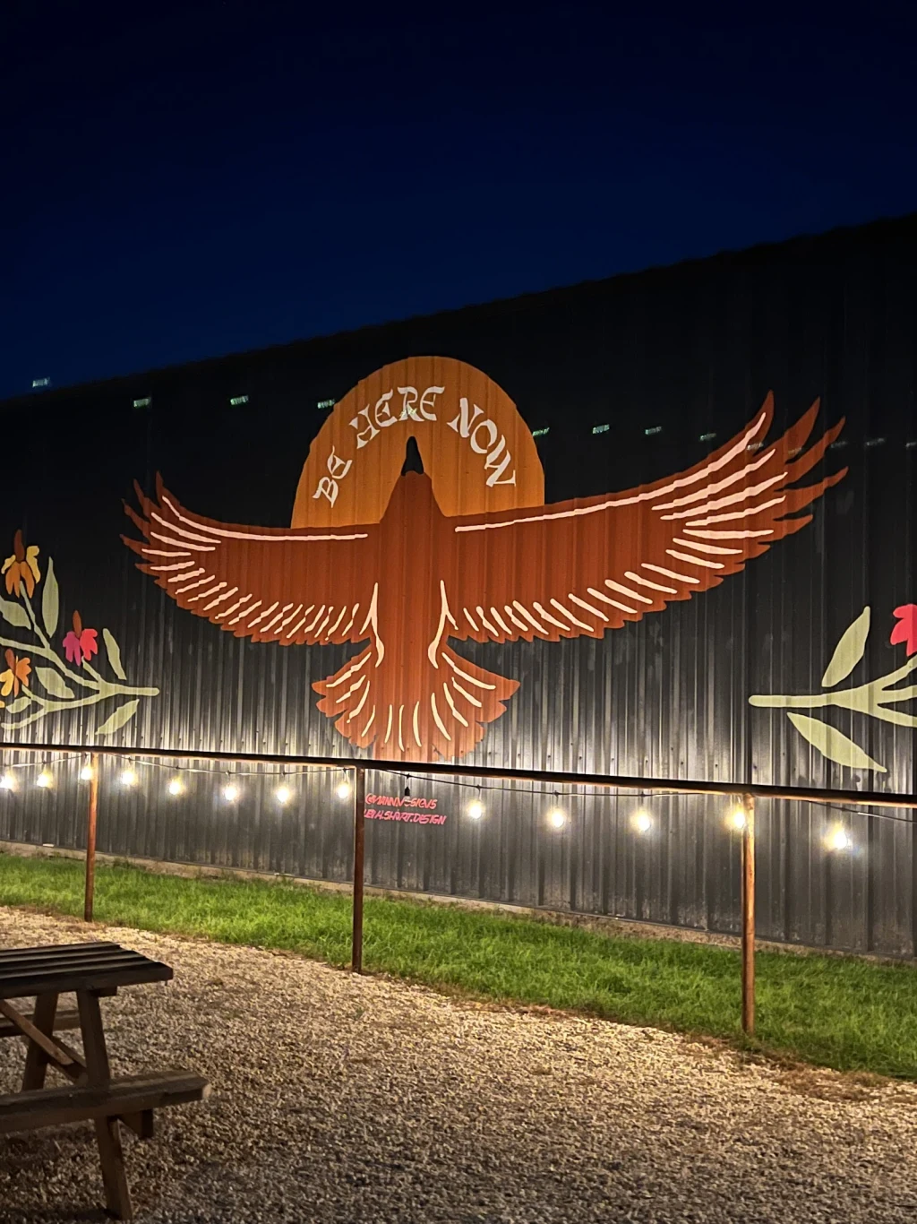 Thunderbird mural with Be Here Now lettering hand-painted across the black metal event barn at Rancho Moonrise, lit by string lights at night